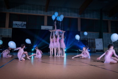 Dziewczynki w różowych strojach. Trzymają białe balony. W środku kadru grupa unosi w górę dziewczynkę z pięcioma balonami. Ta podnosi w górę lewą nogę.