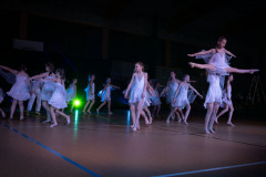 Układ taneczny dziewczyn w białych sukienkach. Na pierwszym planie tancerka podnosi koleżankę robiącą szpagat męski.