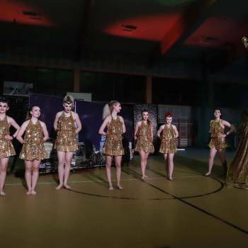Grupa dziewczyn stoi w rzędzie w sali gimnastycznej. Ubrane w złote sukienki. Po prawej dziewczyna na podwyższeniu ukrytym pod zwojami złotego materiału. Unosi w górę prawą rękę.