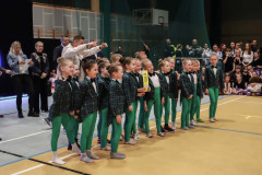 Grupa dziewczynek ubranych w zielone leginsy i marynarki w kratę stoi w dwóch rzędach. Za nimi jurorzy. W głębi kadru publiczność.