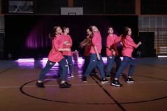 W kole namalowanym na podłodze, oznaczającym środek boiska grupa dziewczyn w układzie tanecznym. Ubrane w różowe koszulki i niebieskie spodnie z dziurami.