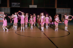 Grupa w trakcie występu. Dziewczynki ubrane w różowe spodenki ogrodniczki, białe koszulki i kolanówki. Włosy związane w dwie kitki.