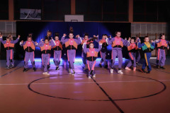 Grupa w trakcie występu w sali sportowej. Na podłodze namalowane koło oznaczające środek boiska. Tancerze ubrani w kolorowe dresy.