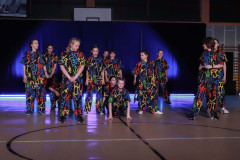 Grupa podczas występu w sali gimnastycznej. Tancerze ubrani w ciemne kostiumy zadrukowane wzorami w kolorach: czerwonym zielonym,. żółtym i niebieskim.