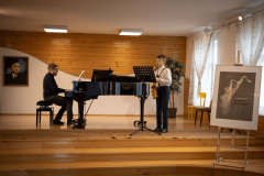 Na scenie, na którą wiodą trzy drewniane stopnie chłopiec gra na saksofonie przed pulpitem, a mężczyzna akompaniuje na fortepianie. W tle portret Karola Szymanowskiego. Po prawej na sztaludze plakat Turnieju Muzycznego.