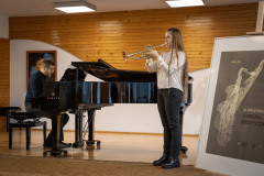 Dziewczyna z bardzo długimi włosami gra na trąbce. Po lewej akompaniujący na fortepianie mężczyzna. Po prawej plakat Turnieju Muzycznego.