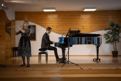 Po lewej grająca na flecie dziewczyna. W środku kadru akompaniujący na fortepianie mężczyzna. W głębi po prawej fikus w donicy.