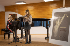 Dziewczyna gra trąbce. W głębi siedząca przy fortepianie kobieta. Z prawej plakat Turnieju Muzycznego.