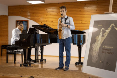 Chłopak gra na saksofonie. Przy fortepianie zasiada mężczyzna. Po prawej plakat Turnieju Muzycznego w antyramie na sztaludze.