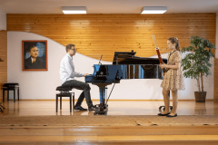 Po prawej dziewczynka w beżowej sukience w kwiatowy wzorek gra na fagocie. Akompaniuje jej mężczyzna na fortepianie. Po lewej wiszący na ścianie portret Karola Szymanowskiego.