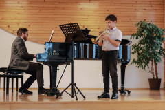 Po prawej chłopiec grający na trąbce. W rogu dorodny fikus. Po lewej mężczyzna akompaniujący na fortepianie.