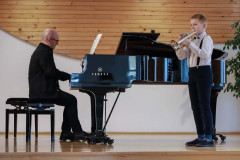 Po prawej chłopiec grający na trąbce. Ubrany w białą koszulę i ciemne spodnie na szelkach. Po prawej akompaniujący na fortepianie mężczyzna.