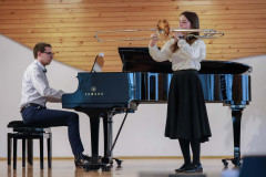 Po prawej dziewczynka grająca na puzonie. Ubrana w białą bluzkę i długą czarną spódnicę. Po lewej mężczyzna akompaniujący na fortepianie. W tle jasna ściana, częściowo pokryta boazerią.