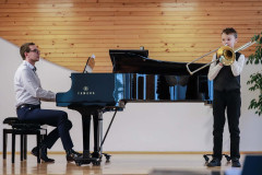 Po prawej chłopiec grający na puzonie. Ubrany w białą koszulę, ciemną kamizelkę i spodnie. Po lewj mężczyzna akompaniujący na fortepianie.