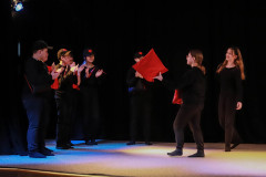 Fragment przedstawienia. Na scenie młodzi aktorzy w czerni. Niektórzy w czapkach z daszkiem. Jedna z dziewczyn trzyma czerwoną poduszkę.