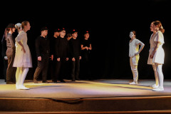 Fragment spektaklu. Na scenie grupa artystów: pośrodku chłopcyu w czerni z czpkami z daszkiem. Po bokach dziewczyny w białych sukienkach.