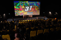 Zdjęcie z górnych rzędów sali. Żółte fotele i siedzący w nich ludzie. W głębi kadru scena, na niej kilka osób i wyświelony slajd 31. Finał WOŚP.