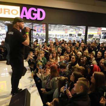 Na skraju sceny stoi mężczyzna w czarnych dresach i koszulce. Na głowie czarny czepek. Sfotografowany z prawego profilu. Przed sceną tłum młodzieży. W tle witryna sklepu Euro AGD.