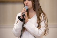 Dłogowłosa dziewczyna w białym swetrze i brązowych spodniach śpiewa do mikrofonu umieszczonego na statywie.