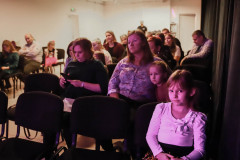 Zdjęcie widowni zasiadającej na krzesłach. W pierwszym rzędzie dziewczynka w jasnej koszulce z kokarda patrzy w obiektyw. Po prawej ciemne kotary.