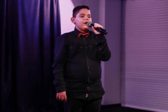 Chłopiec w czerni z czerwoną muszką. W lewej dłoni trzyma mikrofon i śpiewa. W tle wnęka z żaluzją.