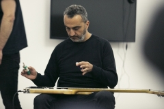 Tony Elieh. W dłoniach trzyma dwa małe urządzenia, które unosi nad gitarą. Instrument spoczywa na jego kolanach.