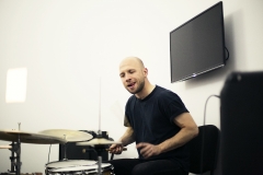 Adam Gołębiewski. Ubrany w czarną koszulkę z krótkimi rękawami. Gra na perkusji. Za jego plecami zamontowany na białej ścianie ekran telewizora.