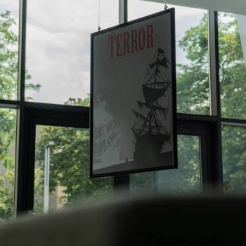 Robione zza kanapy zdjęcie plakatu filmowego Terror, a na nim - czarny i biały żaglowiec. Za szklaną fasadą CKiS widoczna zieleń drzew.