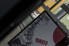 Zrobione ukośnie zdjęcie plakatu filmowego Yamato. Naszkicowany okręt, czerwone litery i czerwone słońce w prawym górnym rogu, zapożyczone z flagi Japonii. Za oknami widoczny blok.