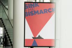 Plakat filmowy Sink The Bismarck. Czerwone, białe i niebieskie trójkąty, czarny okręt. Za plakatem widoczne słynne białe filary Oskardu i schody wiodące do sali widowiskowej.