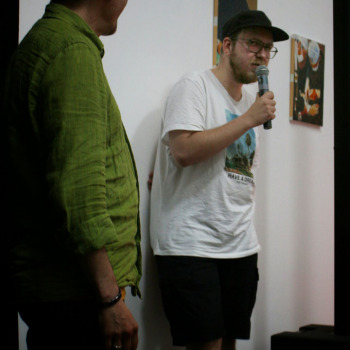 Adam Kempa stoi przy ścianie. W prawej dłoni trzyma mikrofon. Ubrany w białą koszulkę z nadrukiem i czapkę z daszkiem. Obok niego stoi zwrócony bokiem do obiektywu Robert Brzęcki.