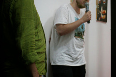Adam Kempa stoi przy ścianie. W prawej dłoni trzyma mikrofon. Ubrany w białą koszulkę z nadrukiem i czapkę z daszkiem. Obok niego stoi zwrócony bokiem do obiektywu Robert Brzęcki.