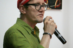Robert Brzęcki w zielonej koszuli i czerwonej czapeczce kolarskiej. W prawej dłoni mikrofon.