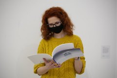 Daria Mach ubrana w żółty sweter ogląda katalog poświęcony artystce. Na twarzy ma czarną maseczkę.