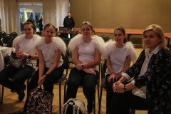 Na krzesłach siedzą cztery dziewczyny w ciemnych spodniach i białych koszulkach. Do ramion doszyte futerko imitujące anielskie skrzydła. Po prawej kobieta w żakiecie w kwiatowy deseń. Patrzą w obiektyw.