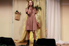 Na scenie dziewczynka w łososiowej sukience z bufkami. Śpiewa do mikrofonu trzymanego w prawej dłoni. Za nią ozdobiony tekstyliami, światełkami i papierową torbą z bombkami parawan.