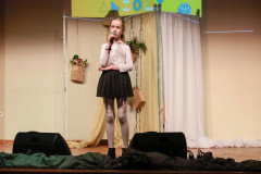 Na scenie dziewczynka w krótkiej czarnej brokatowej spódniczce i białej bluzce. Śpiewa do mikrofonu trzymanego w w prawej dłoni. Za nią ozdobiony tekstyliami, światełkami i papierową torbą z bombkami parawan.