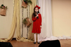 Na scenie dziewczynka w czerwonej sukience z czerwoną kokardą we włosach. Za nią ozdobiony tekstyliami, światełkami i papierową torbą z bombkami parawan.
