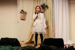 Dziewczynka w białej sukience i białym żakiecie. Śpiewa na scenie do mikrofonu trzymanego w prawej dłoni. Za nią  ozdobiony tekstyliami, światełkami i papierową torbą z bombkami parawan.
