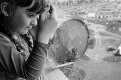 Zdjęcie czarno-białe. Dziewczynka z grzywką wygląda przez okno pokryte kroplami deszczu.