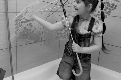 Zdjęcie czarno-białe. Dziewczynka z grzywką i długą kitką stoi w wannie. Trzyma parasol.  Ma na sobie ogrodniczki i kalosze.