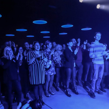 Publiczność na stojąco oklaskuje występ. Nad zgromadzonymi w sali klubowej okrągłe lampy, przypominające świecące spodki.