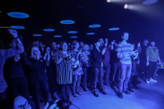 Publiczność na stojąco oklaskuje występ. Nad zgromadzonymi w sali klubowej okrągłe lampy, przypominające świecące spodki.