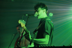Gitarzysta w planie amerykańskim, sfotografowany z lewego profilu. Gra i śpiewa do mikrofonu. W głębi kadru drugi gitarzysta. Za plecami światła reflektora. Zielona poświata.