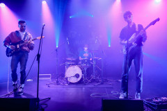 Na pierwszym planie (po prawej i po lewej) dwóch gitarzystów na scenie, w głębi w środku kadru perkusista.