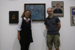 Kobieta (po lewej) i mężczyzna (po prawej) pozują do zdjęcia na tle obrazów wyeksponowanych na ścianie Galerii CKiS Wieża Ciśnień.