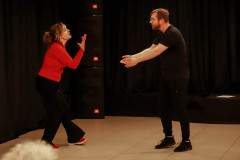 Kobieta w czerwonej bluzce, czerwonych butach sportowych i czarnych dzwonach gestykuluje. Naprzeciwko niej mężczyzna w czerni i okularach. Wyciąga ręce,