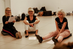 Trzy kobiety siedzą na podłodze na szarych siedziskach. Śmieją się. Przy ścianie torby i buty.