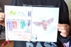 Uśmiechnięty chłopiec trzyma w dłoniach rysunek. Po lewej stronie otwarta szafa z wiszącymi na wieszakach ubraniami. Po prawej owad.