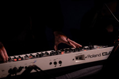 Zbliżenie na dłonie na keyboardzie z napisem Roland. Pozostała część zdjęcia w ciemnościach.
